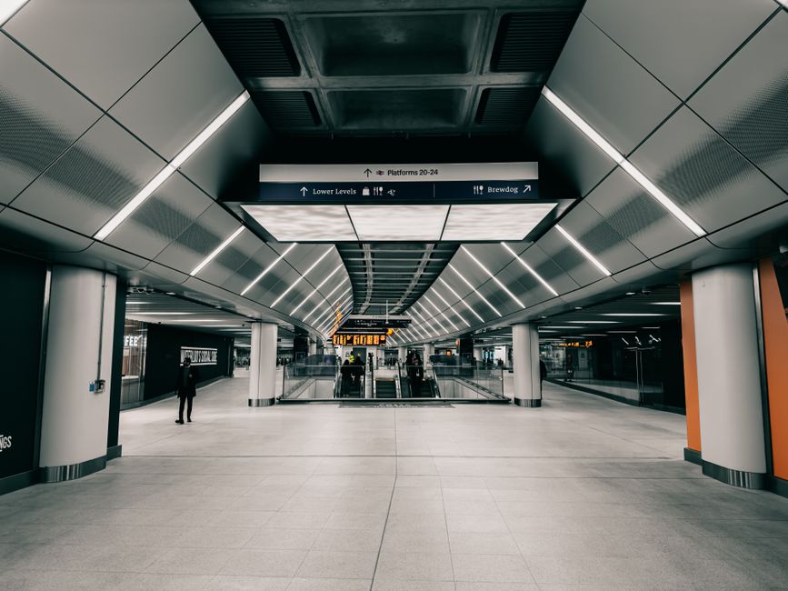 A spacious, modern underground station with wide, clean ceramic tiled floors and sleek, metallic ceiling panels featuring integrated LED lighting. The station has a symmetrical layout with large white