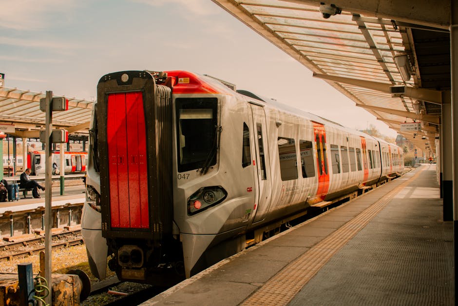 A spacious, modern underground station with wide, clean ceramic tiled floors and sleek, metallic ceiling panels featuring integrated LED lighting. The station has a symmetrical layout with large white